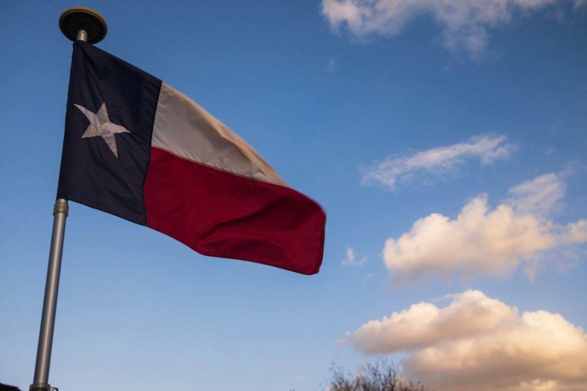 Bandera del estado de Texas ondeando en un mástil metálico contra un cielo azul con nubes blancas.